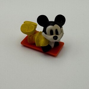 May include: A small, vintage Mickey Mouse figurine in a reclining pose. The figure is wearing yellow shorts and shoes, with a white shirt and black ears. It rests on a small, orange rectangular base.