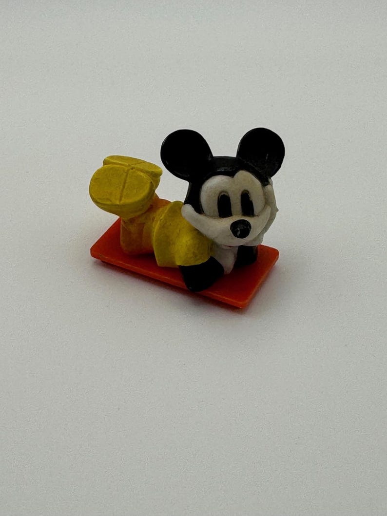 May include: A small, vintage Mickey Mouse figurine in a reclining pose. The figure is wearing yellow shorts and shoes, with a white shirt and black ears. It rests on a small, orange rectangular base.