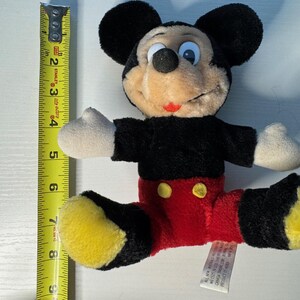 May include: A plush Mickey Mouse toy, with black ears, a black torso, red shorts, and yellow shoes. The toy is approximately 20 cm long. The toy has a tag attached. A yellow measuring tape is next to the toy.