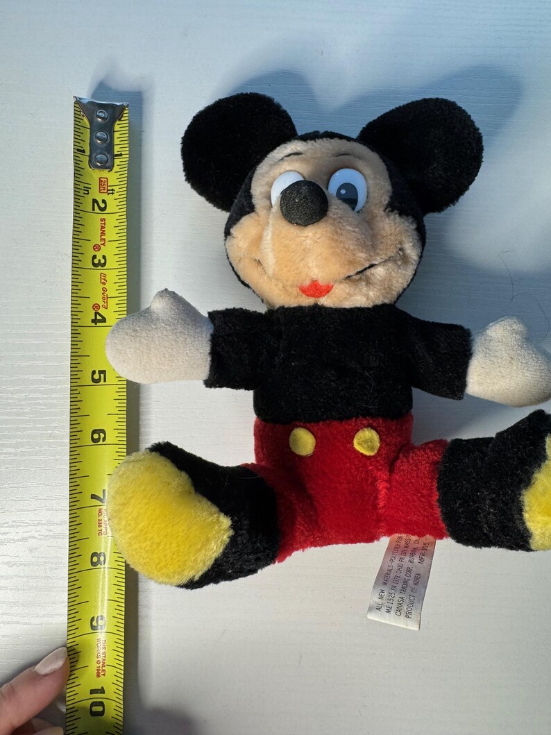 May include: A plush Mickey Mouse toy, with black ears, a black torso, red shorts, and yellow shoes. The toy is approximately 20 cm long. The toy has a tag attached. A yellow measuring tape is next to the toy.