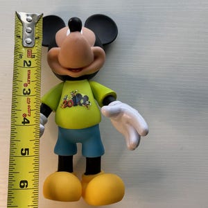 May include: A Mickey Mouse figurine, approximately 6 inches tall, wearing a yellow shirt with a graphic, blue shorts, and yellow shoes. The figure has black ears, a tan face, and white gloves. A measuring tape is next to the figure.