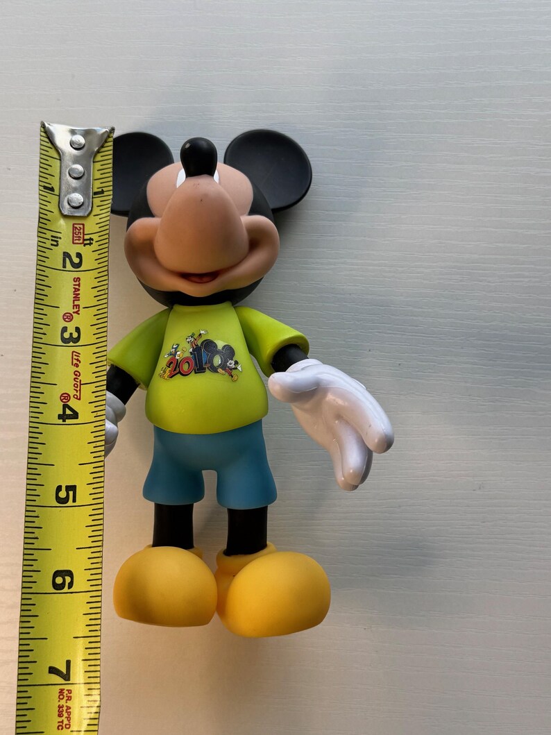 May include: A Mickey Mouse figurine, approximately 6 inches tall, wearing a yellow shirt with a graphic, blue shorts, and yellow shoes. The figure has black ears, a tan face, and white gloves. A measuring tape is next to the figure.