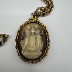 May include: Vintage gold-tone cameo pendant necklace. The oval pendant features a relief carving of two figures in cream against a brown background. The pendant is framed in a decorative gold setting and hangs from a gold chain.