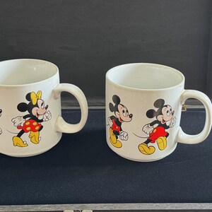 May include: Two white ceramic mugs with handles. One mug features Mickey Mouse and Minnie Mouse in red and black outfits. The other mug features Mickey Mouse and Minnie Mouse in black and red outfits.