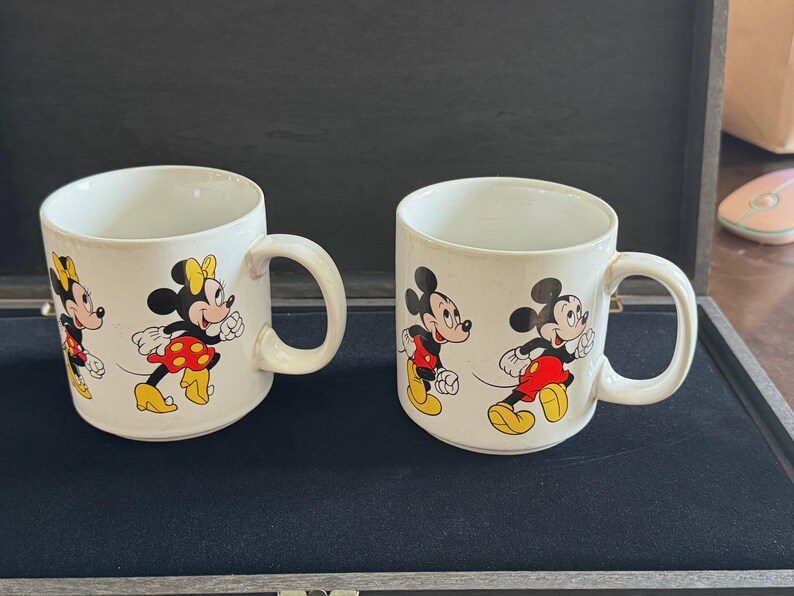 May include: Two white ceramic mugs with handles. One mug features Mickey Mouse and Minnie Mouse in red and black outfits. The other mug features Mickey Mouse and Minnie Mouse in black and red outfits.