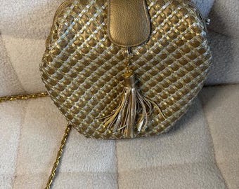 May include: Gold-colored woven handbag with a chain strap and a tassel. The bag has a flap closure and a textured pattern. The chain strap is also gold. The bag is sitting on a textured surface.