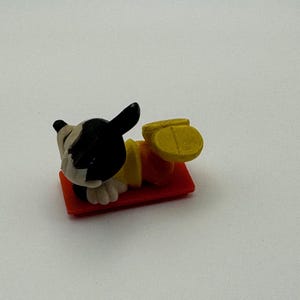 May include: A small, plastic figurine of a dog lying on a red rectangular base. The dog has a black and white head, a yellow shirt, and orange pants. The dog's feet are yellow. The figurine is a toy or collectible item.