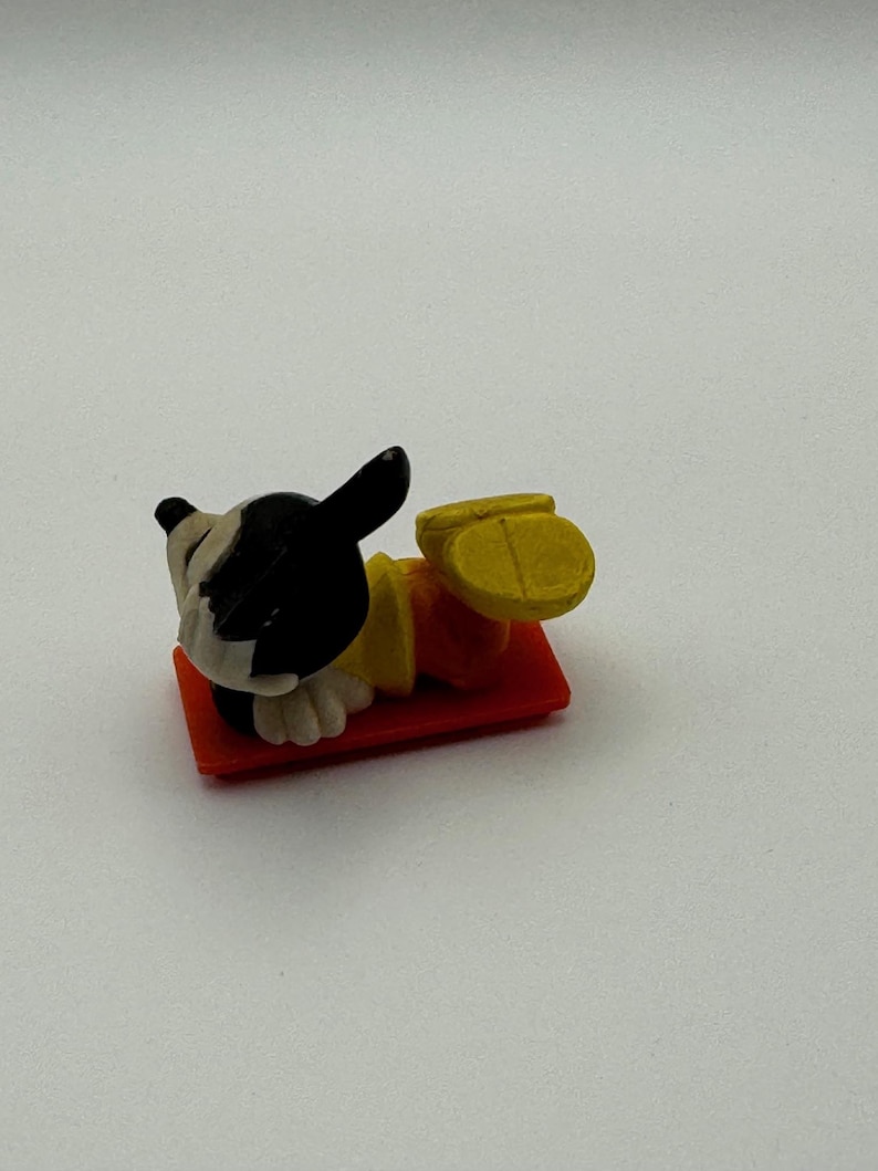 May include: A small, plastic figurine of a dog lying on a red rectangular base. The dog has a black and white head, a yellow shirt, and orange pants. The dog's feet are yellow. The figurine is a toy or collectible item.
