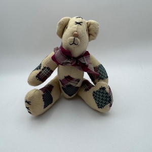 May include: A beige teddy bear with a burgundy plaid bow tie. The bear has patchwork accents on its arms and legs in shades of green, burgundy, and navy. The bear is sitting with its legs spread.