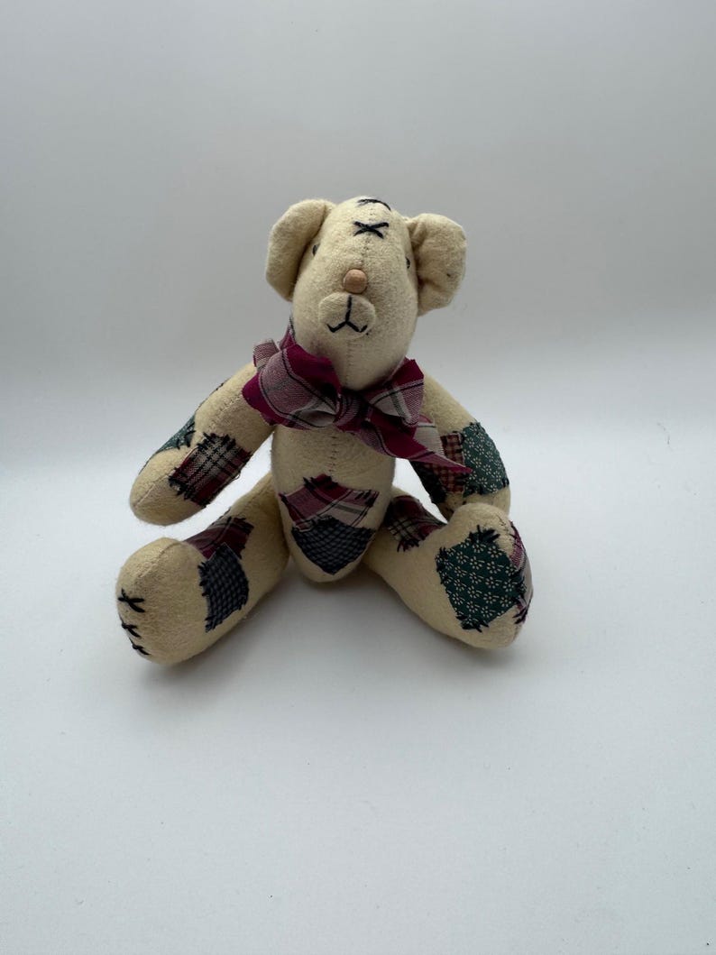 May include: A beige teddy bear with a burgundy plaid bow tie. The bear has patchwork accents on its arms and legs in shades of green, burgundy, and navy. The bear is sitting with its legs spread.