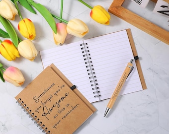May include: A spiral-bound notebook with the text "Sometimes you forget you're Awesome so this is your reminder." A wooden pen and a wooden framed calendar are also present. Yellow and pink tulips are arranged around the notebook.