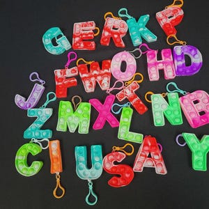 May include: A collection of colorful alphabet letter keychains in various colors, including red, green, blue, and purple. Each letter features a bubble-popping design and a metal clasp for attachment. The letters are arranged on a black background.