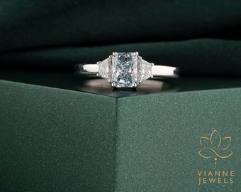 May include: A silver ring featuring a rectangular, light blue gemstone flanked by two triangular, clear gemstones. The ring is set against a dark green backdrop, with the brand name "VIANNE JEWELS" visible in the lower right corner.