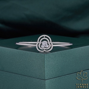 May include: A silver bangle bracelet with a flower-shaped design featuring a clear, faceted gemstone in the centre. The bracelet is on a green surface.
