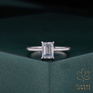 May include: A silver engagement ring with an emerald-cut diamond. The ring is set on a dark green surface. The ring's band is thin and delicate, and the diamond is the focal point. The ring is a classic and elegant design.