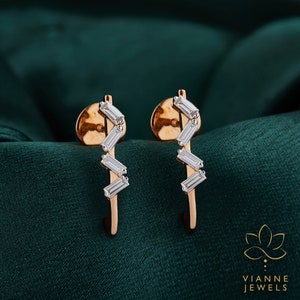 May include: A pair of rose gold earrings with a zig-zag design featuring small, clear baguette-shaped diamonds. The earrings are set against a dark green background.