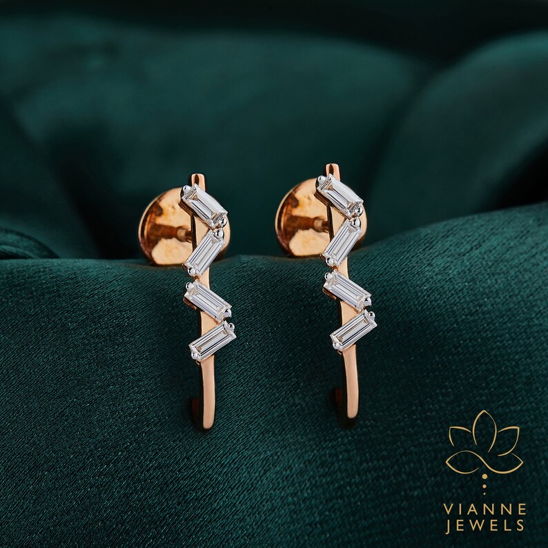 May include: A pair of rose gold earrings with a zig-zag design featuring small, clear baguette-shaped diamonds. The earrings are set against a dark green background.