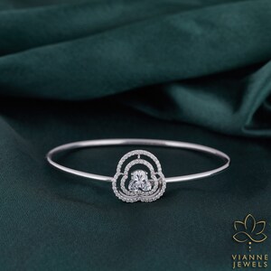 May include: A silver bangle bracelet with a delicate, cloud-shaped design featuring a large, clear, oval-shaped gemstone surrounded by smaller, round gemstones.