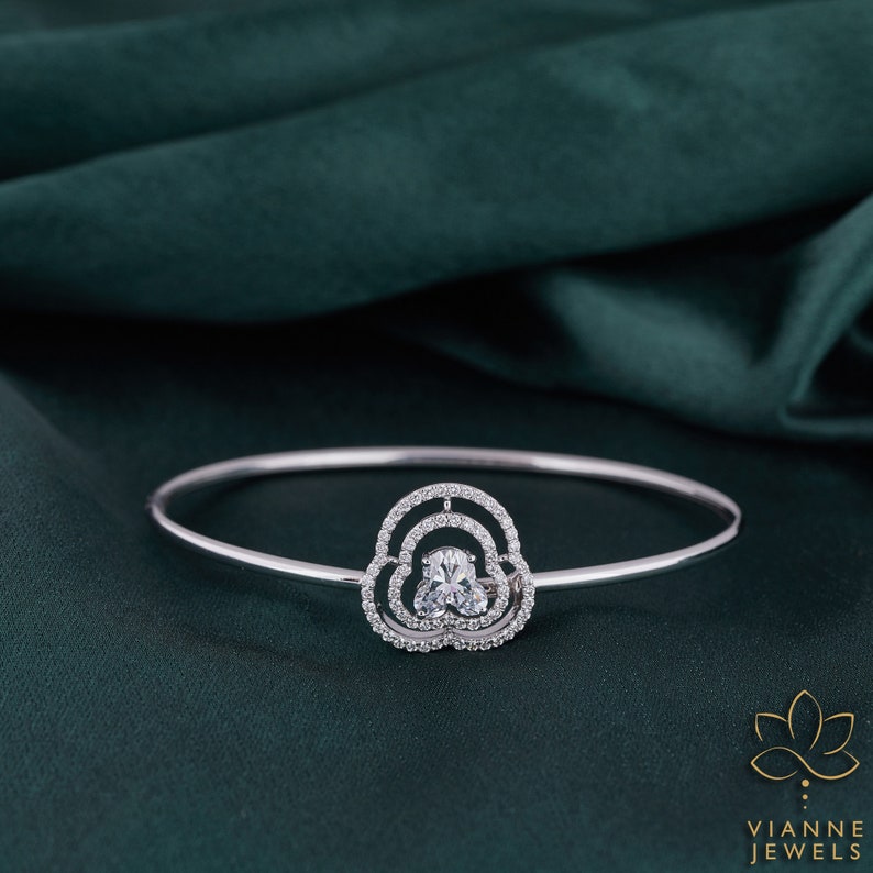 May include: A silver bangle bracelet with a delicate, cloud-shaped design featuring a large, clear, oval-shaped gemstone surrounded by smaller, round gemstones.