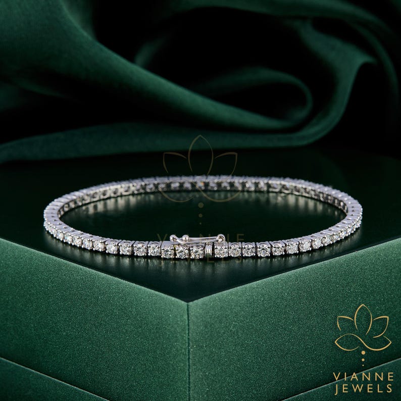 May include: A tennis bracelet with numerous small, round, clear gemstones set in a silver-colored metal. The bracelet is displayed on a dark green surface. The clasp is visible. The brand name "VIANNE JEWELS" is in the lower right corner.
