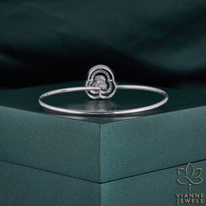 May include: A silver bangle bracelet featuring a large, round, clear gemstone set in a silver setting. The gemstone is surrounded by smaller, clear gemstones. The bracelet is displayed on a green velvet box.