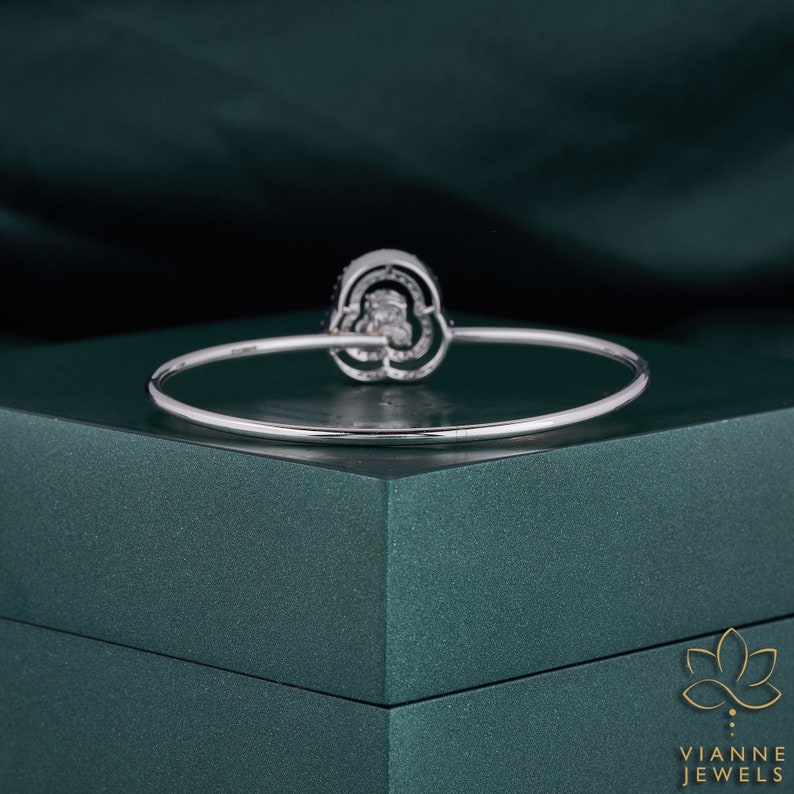 May include: A silver bangle bracelet featuring a large, round, clear gemstone set in a silver setting. The gemstone is surrounded by smaller, clear gemstones. The bracelet is displayed on a green velvet box.