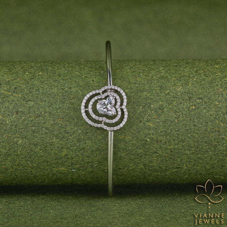 May include: A silver bangle bracelet with a delicate, flower-shaped design featuring a single, clear gemstone in the centre. The bracelet is set against a green background.