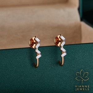 May include: A pair of gold hoop earrings with a row of baguette-shaped diamonds set in a staggered pattern. The earrings are on a green surface.