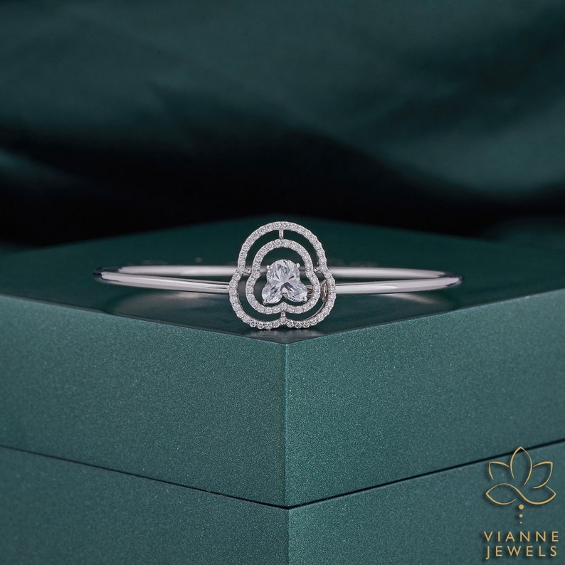 May include: A silver bangle bracelet with a flower-shaped design featuring a clear, faceted gemstone in the centre. The bracelet is on a green surface.