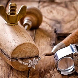 May include: A diamond ring with a floral design is held in a wooden ring clamp. A magnifying glass and other jewelry tools are on a wooden surface. The ring has a silver band and a cluster of diamonds.