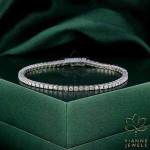 May include: A delicate silver bracelet with numerous small, sparkling gemstones. The bracelet is set against a dark green backdrop, highlighting its brilliance. The bracelet is a tennis style, perfect for adding a touch of elegance to any outfit.