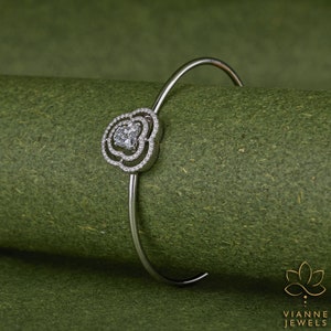 May include: A silver bangle bracelet with a heart-shaped design featuring a large clear stone surrounded by smaller clear stones.