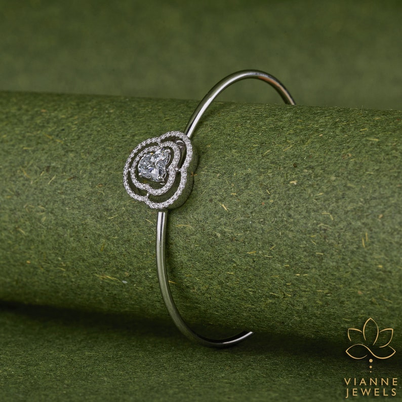 May include: A silver bangle bracelet with a heart-shaped design featuring a large clear stone surrounded by smaller clear stones.