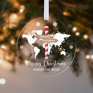 May include: A clear, circular Christmas ornament with a world map design and the text "Merry Christmas Across the Miles." It features a red and white striped pole with directional signs pointing to England and Australia. Includes snowflake accents and a white ribbon.