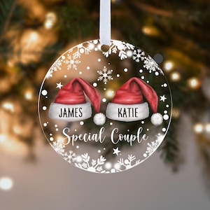 May include: A clear, circular Christmas bauble with a white ribbon. It displays two red Santa hats, each with a name: "JAMES" and "KATIE". Below, the words "Special Couple" are written, surrounded by snowflakes and stars.