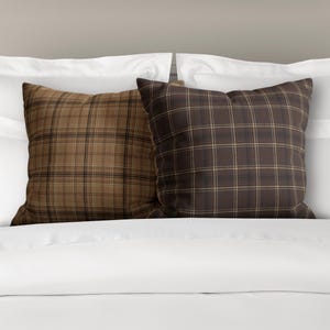 May include: Two square throw pillows with a brown and tan plaid pattern. The pillows are on a white bed.