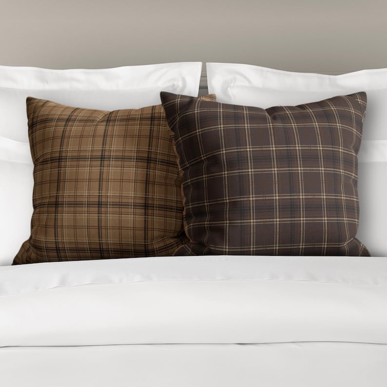 May include: Two square throw pillows with a brown and tan plaid pattern. The pillows are on a white bed.