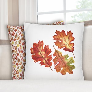 May include: White square throw pillow featuring three watercolor oak leaves in shades of red, orange, and yellow. A second pillow with a repeating pattern of autumn leaves is visible. The pillows are on a white surface.
