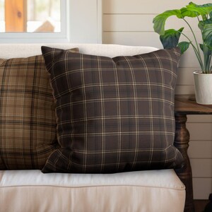 May include: Two brown and tan plaid throw pillows on a cream colored couch. The pillows are made of a soft, woven fabric and have a subtle sheen.