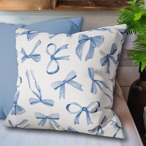 May include: A white pillow with a blue watercolor bow pattern. The pillow is on a light blue pillow and a light brown surface.