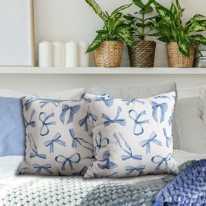 May include: Two decorative throw pillows with a white background and blue watercolor bow patterns. The pillows are on a white couch with a blue blanket.
