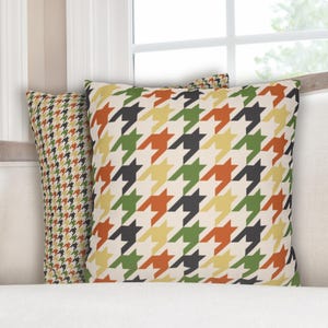 May include: Two decorative throw pillows with a houndstooth pattern in various colors, including black, green, orange, and yellow, on a cream background. The pillows are placed on a white sofa near a window.