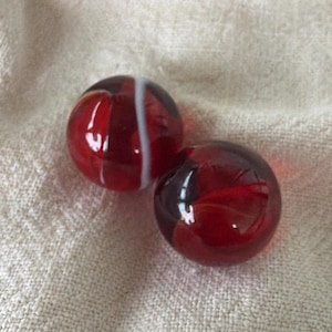 May include: Two spherical glass marbles featuring a deep red base color with swirling dark patterns. One marble displays a distinct vertical white stripe running across its surface, while the other shows a smooth, glossy finish with internal red hues.