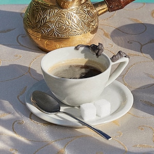 May include: A white ceramic coffee cup with a dark brown coffee inside. The cup has a decorative handle and is sitting on a white saucer with two sugar cubes. A silver spoon is on the saucer.