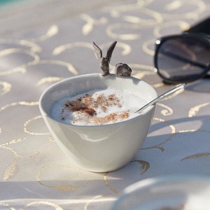 May include: A white ceramic bowl filled with a creamy white beverage topped with a sprinkle of brown powder. A small gray ceramic figurine of a bird sits on the rim of the bowl.