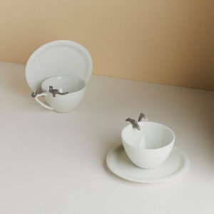 Może przedstawiać: Dwie białe ceramiczne filiżanki do herbaty z pasującymi spodkami. Jedna filiżanka ma figurkę delfina na uchwycie, druga ma figurkę ptaka na uchwycie.