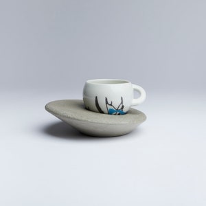Könnte beinhalten: Eine weiße Keramiktasse mit einem abstrakten Design in Blau und Schwarz steht auf einer grauen Keramiksaucers.