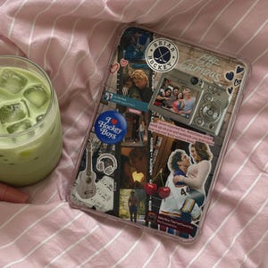 Könnte beinhalten: Ein klares Glas gefüllt mit hellgrünem Matcha-Latte und Eiswürfeln, mit einem Glasstrohhalm, der einen kleinen braunen Bärenanhänger hat. Daneben liegen eine rosa Lippenpflege-Tube und eine rechteckige Tablethülle, die mit Stickern beklebt ist.