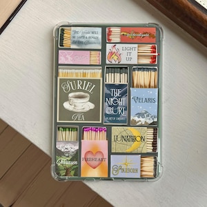 Könnte beinhalten: Eine transparente Handyhülle gefüllt mit Streichholzschachteln. Die Schachteln zeigen verschiedene Designs und Texte, darunter "THE WORLD WILL BE SAVED & REMADE BY THE DREAMERS", "LIGHT IT UP" und "JURIEL TEA". Weitere Designs sind eine Berglandschaft und ein Herz.