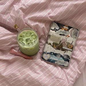 May include: A glass of iced matcha latte with a bear-topped straw, a pink Rhode lip treatment tube, and a rectangular tablet case featuring a collage of fantasy-themed imagery and text including "Through love, all is possible" and "Hello, Feyre Darling."